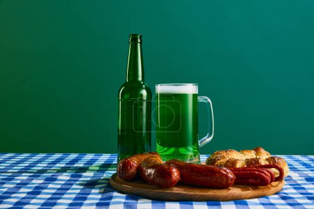 Téléchargez les photos : Mug with foamy green beer and plate of grilled sausages and buns on checkered tablecloth on green background. Concept of st patricks day celebration, traditions, alcohol drinks, taste, Irish holiday - en image libre de droit