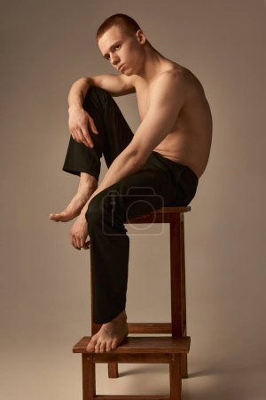 Photo for Serious. Portrait of handsome young boy with short red hair posing on chair shirtless in pants over light brown studio background. Concept of mens health and beauty, body care, fitness. Body art - Royalty Free Image
