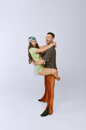 Téléchargez les photos : Smiling. Happy young man and woman in stylish retro outfits dancing disco dance isolated over grey studio background. 70s fashion, hobby, creativity, hippie lifestyle, American culture - en image libre de droit