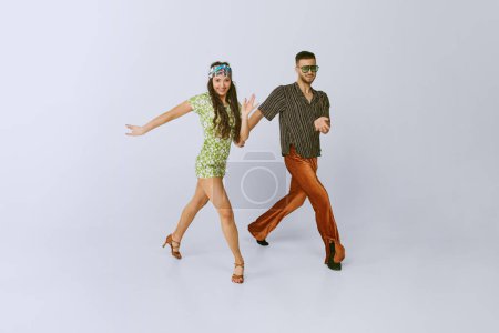 Téléchargez les photos : Coordinated steps. Attractive young couple, man and woman in retro clothes dancing disco isolated over grey studio background. 70s fashion, hobby, creativity, hippie lifestyle, American culture - en image libre de droit
