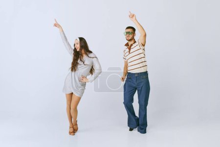 Téléchargez les photos : Having fun. Young beautiful girl in silver dress and stylish man dancing disco dance isolated over grey studio background. 70s fashion, hobby, creativity, hippie lifestyle, American culture - en image libre de droit