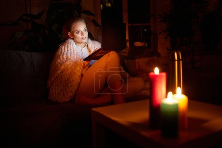 Photo for Adjusting to unstable electricity. Young girl, freelancer sitting on sofa at home in evening and working, watching movie on laptop with candle light. Blackout. Concept of power outage - Royalty Free Image