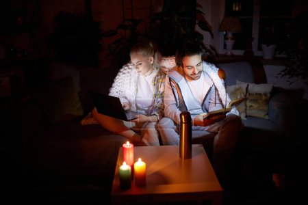 Photo for Cozy family evening. No electricity. Young man and woman sitting on sofa at home in evening, working on laptop, reading book with candles light. Blackout. Concept of power outage, relationship - Royalty Free Image