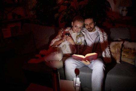 Photo for Lovely couple. Cozy evening. Man and woman in warm blanket sitting on sofa, hugging and reading book together. No electricity, blackout. Concept of power outage, relationship, lifestyle - Royalty Free Image
