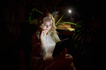 Photo for Young beautiful woman sitting in dark room sitting on chair and reading book on laptop at home in the evening with battery powered lamp light. No electricity, blackout. Concept of power outage - Royalty Free Image