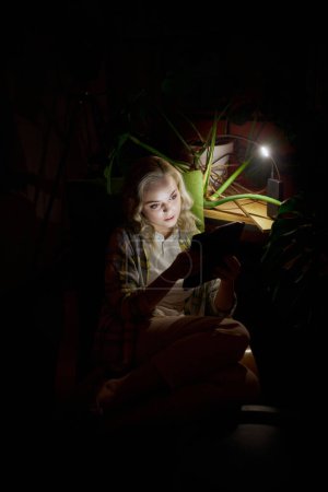 Foto de Joven hermosa mujer sentada en habitación oscura sentada en la silla con el ordenador portátil en casa por la noche con la luz de la lámpara alimentado por batería. Sin electricidad, apagón. Concepto de corte de energía, trabajo independiente - Imagen libre de derechos