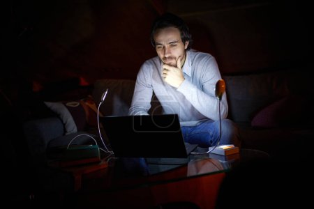 Foto de Ajustándose a la electricidad inestable. Hombre, freelancer sentado en el sofá en casa por la noche y trabajando en el ordenador portátil con la luz de la lámpara de batería. Apagón, sin electricidad. Concepto de corte de energía - Imagen libre de derechos