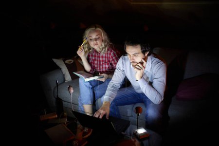 Photo for Man and woman, couple sitting on sofa at home in the evening and studying, working with little light of battery lamp. No electricity, blackout. Concept of power outage, online education, freelance - Royalty Free Image