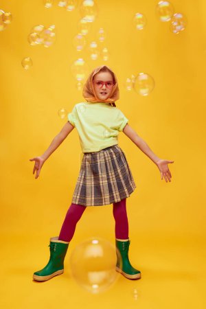 Téléchargez les photos : Beautiful little girl, child in stylish clothes, skirt and rubber boots posing on yellow studio background with soap bubbles. Concept of childhood, emotions, fun, fashion, lifestyle, facial expression - en image libre de droit