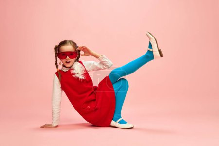 Téléchargez les photos : Feeling comfortable. Girl, child in red dress, blue tights and red glasses posing over pink studio background. Concept of childhood, emotions, fun, fashion, lifestyle, facial expression - en image libre de droit