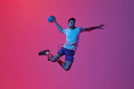 Téléchargez les photos : Lancer la balle dans le saut. Jeune homme, entraînement professionnel de joueur de handball, jouant isolé sur fond rose dégradé sous la lumière du néon. Concept de sport, action, mouvement, championnat, mode de vie sportif - en image libre de droit