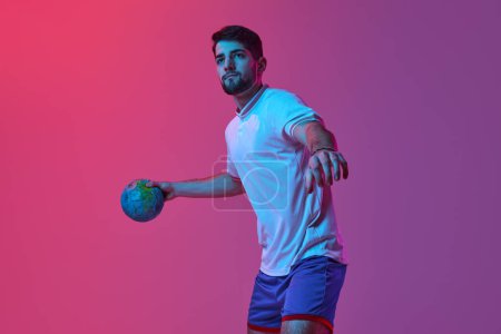 Photo for High concentration. Young man, professional handball player training, playing isolated over gradient pink background in neon light. Concept of sport, action, motion, championship, sportive lifestyle - Royalty Free Image