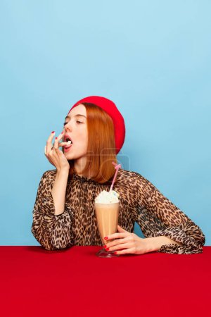 Téléchargez les photos : Tasting whipped cream. Yummy. Young woman in red beret drinking milkshake over blue studio background. Food pop art photography. Complementary colors. Concept of art, beauty, food. Copy space for ad - en image libre de droit