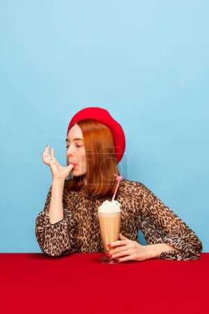 Téléchargez les photos : Douce vie. Jeune femme au béret rouge dégustant de délicieux milkshake à la crème fouettée sur fond bleu studio. Photographie pop art culinaire. Couleurs complémentaires. Concept d'art, de beauté, de nourriture. Publicité - en image libre de droit