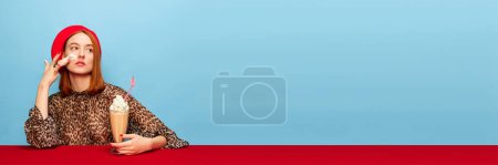 Foto de Natural ingredients. Young woman in red beret tasting milkshake, applying whipped cream over blue studio background. Food pop art photography. Complementary colors. Concept of art, beauty, food. - Imagen libre de derechos