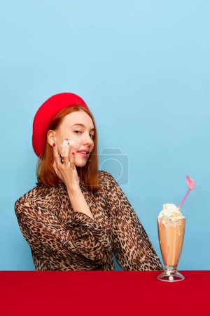 Téléchargez les photos : Appliquer de la crème fouettée sur le visage. Jeune femme au béret rouge, avec milkshake sur fond bleu studio. Photographie pop art culinaire. Couleurs complémentaires. Concept d'art, de beauté, de nourriture. Espace de copie pour la publicité - en image libre de droit