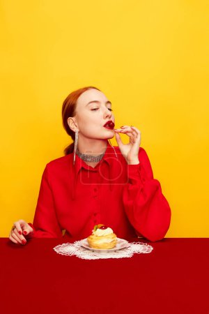Téléchargez les photos : Beautiful young girl in red shirt tasting delicious cake with cream filling on yellow studio background. Food pop art photography. Complementary colors. Concept of art, beauty, food. Copy space for ad - en image libre de droit