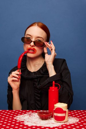 Photo for Young beautiful girl in trendy sunglasses eating spicy red chilli pepper and mayonnaise over blue background. Food pop art photography. Complementary colors. Concept of art, beauty, food. - Royalty Free Image