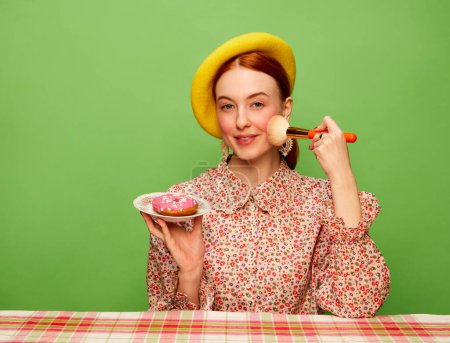 Foto de Beautiful girl with blushing cheeks eating pink donut over green background. Countryside style. Food pop art photography. Complementary colors. Concept of art, beauty, food. Copy space for ad, text. - Imagen libre de derechos