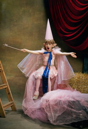 Photo for Emotional little kid, beautiful girl in image of fairy magical elf posing in cute pink dress over vintage circus background. Retro circus performance. Concept of dreams, art, vintage style, childhood - Royalty Free Image