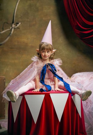 Foto de Magia. Emociones. Niña linda, niño en la imagen de duende mágico posando en vestido rosa sobre el fondo oscuro del circo entre bastidores. Actuación en circo retro. Concepto de sueños, arte, estilo vintage, infancia - Imagen libre de derechos