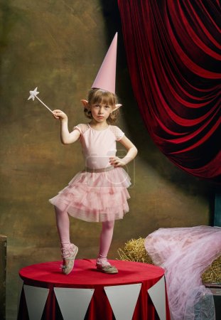 Téléchargez les photos : Magic. Little cute girl, kid in image of magical elf posing in pink dress over dark circus backstage background. Retro circus performance. Concept of dreams, art, fashion, vintage style. - en image libre de droit