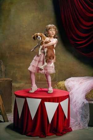 Téléchargez les photos : Magique. Petite fille mignonne, enfant à l'image d'elfe magique posant avec chien sur fond de coulisses de cirque sombre. Spectacle de cirque rétro. Concept de rêve, art, mode, style vintage, enfance - en image libre de droit