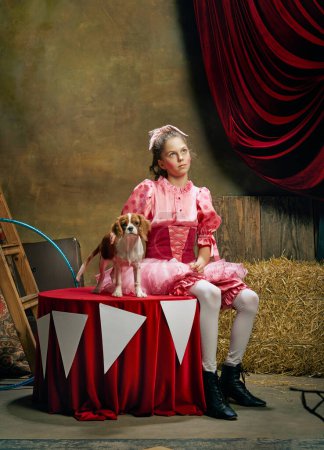 Foto de Hermosa niña en vestido rosa festivo posando con lindo perro tranquilo sobre fondo oscuro circo vintage. Concepto de circo retro, vacaciones, sueños, arte, moda, estilo vintage - Imagen libre de derechos
