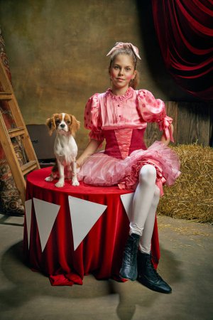 Téléchargez les photos : Belle petite fille en robe rose festive posant avec un chien mignon intelligent sur fond de cirque vintage sombre. Concept de cirque rétro, vacances, rêves, art, mode, style vintage - en image libre de droit