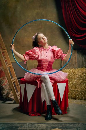 Téléchargez les photos : Belle petite fille en robe rose festive posant avec cerceau gymnastique sur fond de cirque vintage sombre. Concept de cirque rétro, vacances, rêves, art, mode, style vintage - en image libre de droit