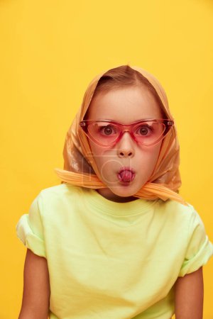 Téléchargez les photos : Grimacing, funny face. Beautiful playful little girl, child in stylish clothes posing over yellow studio background. Concept of childhood, emotions, fun, fashion, lifestyle, facial expression - en image libre de droit