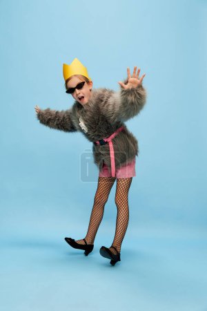 Photo for Balancing. Beautiful little girl, child in trendy fur coat, paper crown and sunglasses posing over blue studio background. Concept of childhood, emotions, fun, fashion, lifestyle, facial expression - Royalty Free Image