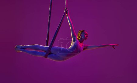 Téléchargez les photos : Un regard gracieux. Jeune fille flexible faisant du yoga aérien, s'entraînant sur fond de studio violet sous la lumière du néon. Concept de fitness, mode de vie sportif, santé, force, yoga aérien, yoga anti-gravité - en image libre de droit