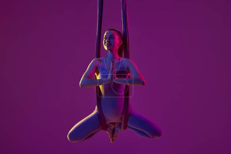Foto de Yoga pose, meditation and relaxation. Young flexible girl doing aerial yoga, training over purple studio background in neon light. Concept of fitness, sportive lifestyle, healthcare, strength - Imagen libre de derechos