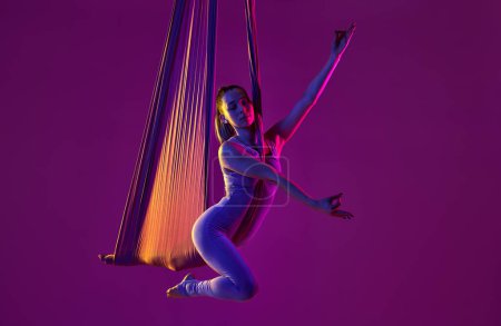 Téléchargez les photos : Garder l'équilibre intérieur. Jeune fille flexible faisant du yoga aérien, formation sur fond de studio violet dans la lumière au néon. Concept de fitness, mode de vie sportif, santé, force, yoga aérien, yoga anti-gravité - en image libre de droit