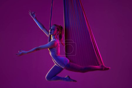Foto de Vibraciones positivas y relajación. Joven chica flexible haciendo yoga aéreo, entrenamiento sobre fondo estudio púrpura en luz de neón. Concepto de fitness, salud, fuerza, yoga aéreo, yoga anti-gravedad - Imagen libre de derechos