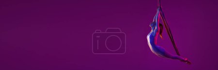 Téléchargez les photos : Jeune fille flexible faisant du yoga aérien, s'entraînant sur fond de studio violet sous la lumière du néon. Concept de fitness, santé, force, yoga aérien, yoga anti-gravité. Bannière, flyer. Espace de copie pour la publicité - en image libre de droit