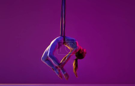 Foto de Stretching in air. Young flexible girl doing aerial yoga, training over purple studio background in neon light. Concept of fitness, sportive lifestyle, health, strength, aerial yoga, anti-gravity yoga - Imagen libre de derechos