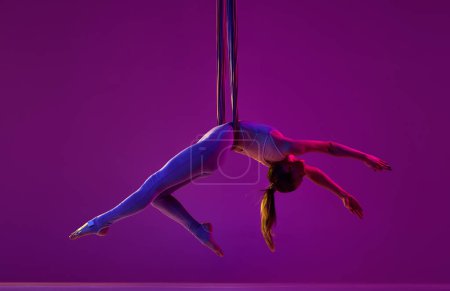 Foto de Relajante. Joven chica flexible haciendo yoga con mosca aérea, entrenamiento sobre fondo estudio púrpura en luz de neón. Concepto de fitness, estilo de vida deportivo, salud, fuerza, yoga aéreo, yoga anti-gravedad - Imagen libre de derechos