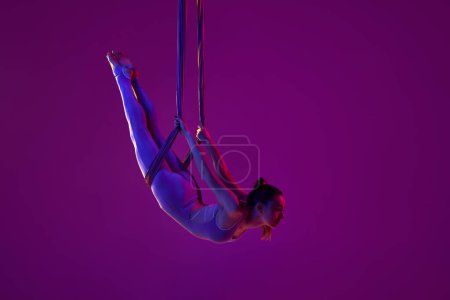 Foto de Strong body. Young flexible girl doing aerial yoga, training over purple studio background in neon light. Concept of fitness, sportive lifestyle, health, strength, aerial yoga, anti-gravity yoga - Imagen libre de derechos