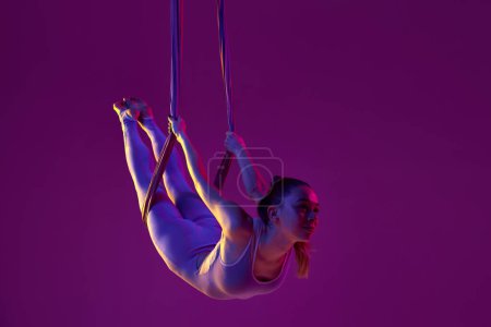 Foto de Relaxing activity. Young flexible girl doing aerial yoga, training over purple studio background in neon light. Concept of fitness, sportive lifestyle, health, strength, aerial yoga, anti-gravity yoga - Imagen libre de derechos