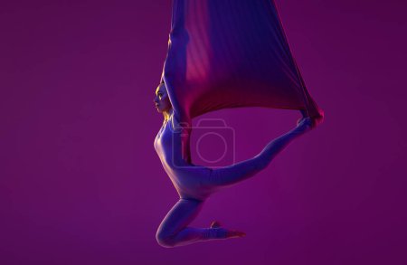 Foto de Like a bat. Young flexible girl doing aerial yoga, training over purple studio background in neon light. Concept of fitness, sportive lifestyle, health, strength, aerial yoga, anti-gravity yoga - Imagen libre de derechos