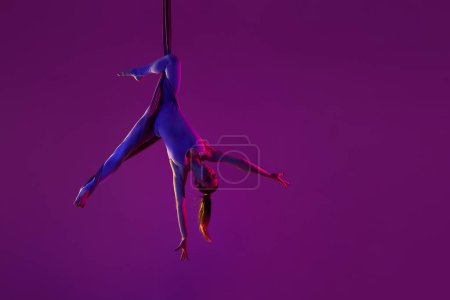 Téléchargez les photos : Ça tourne. Jeune fille flexible faisant du yoga aérien, s'entraînant sur fond de studio violet sous la lumière du néon. Concept de fitness, mode de vie sportif, santé, force, yoga aérien, yoga anti-gravité - en image libre de droit