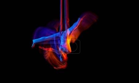 Photo for Twine. Young flexible girl doing aerial yoga, training over black studio background in neon with mixed lights. Concept of fitness, sportive lifestyle, health, strength, aerial yoga, anti-gravity yoga - Royalty Free Image
