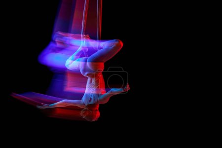 Téléchargez les photos : Balance. Young flexible girl doing aerial yoga, training on black studio background in neon with mixed lights. Concept of fitness, sportive lifestyle, health, strength, aerial yoga, anti-gravity yoga - en image libre de droit