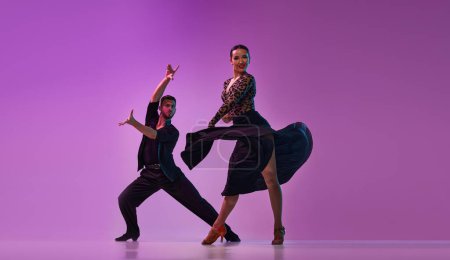 Téléchargez les photos : Artistic young man and woman, professional dancers in stylish stage costumes performing tango over purple background on neon lights. Concept of lifestyle, beauty of movements, emotions, fashion, art - en image libre de droit