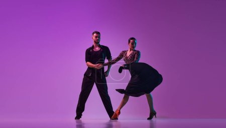 Photo for Artistic young man and woman, professional dancers in stylish stage costumes performing tango over purple background on neon lights. Concept of lifestyle, beauty of movements, emotions, fashion, art - Royalty Free Image