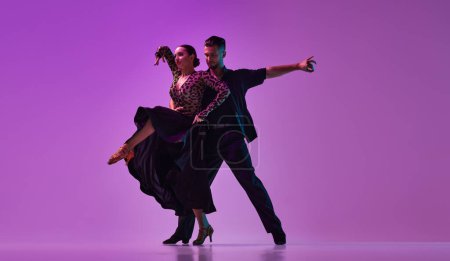 Téléchargez les photos : Talented young man and woman, professional dancers performing tango over purple background on neon lights. Concept of hobby, lifestyle, action, beauty of movements, emotions, fashion, art - en image libre de droit