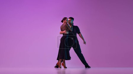 Foto de Joven talentoso hombre y mujer, bailarines profesionales con trajes elegantes realizando tango sobre fondo púrpura sobre luces de neón. Concepto de, estilo de vida, acción, belleza de movimientos, emociones, moda, arte - Imagen libre de derechos