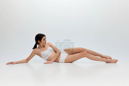 Photo for Self-care. Portrait of young beautiful girl with slim body posing in white cotton underwear posing over grey studio background. Concept of body and skin care, fitness, natural beauty, health, wellness - Royalty Free Image